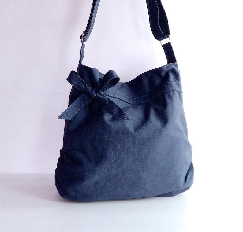 May include: A dark blue canvas shoulder bag with a large bow on the front. The bag has a long adjustable strap.