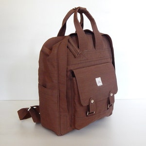 Pode incluir: Uma mochila de lona marrom com um bolso frontal grande e duas alças ajustáveis. A mochila tem um pequeno rótulo branco com o texto "Baby K's" nela.