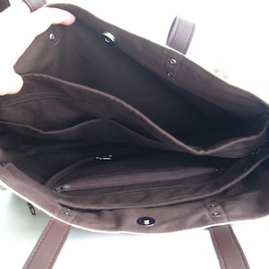 K&ouml;nnte beinhalten: Beige Tragetasche mit braunem Innenraum und Griffen. Die Tasche hat mehrere Innentaschen, darunter Rei&szlig;verschlussf&auml;cher. Die Tasche ist offen und zeigt die Innenorganisation. Die Griffe sind dunkelbraun.