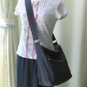 May include: A dark gray shoulder bag with a long strap. The bag has a flap with a zipper closure and a small pocket on the front. The bag is being worn by a person wearing a white shirt with a black and red pattern.