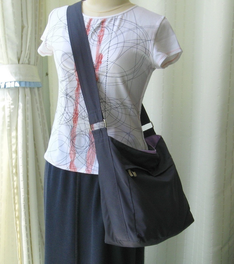 May include: A dark gray shoulder bag with a long strap. The bag has a flap with a zipper closure and a small pocket on the front. The bag is being worn by a person wearing a white shirt with a black and red pattern.