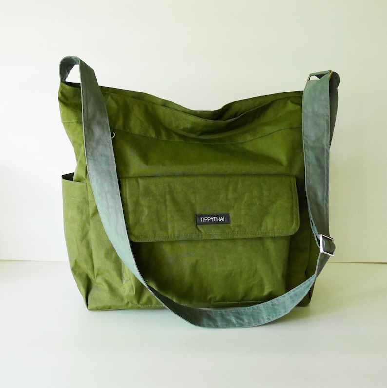 K&ouml;nnte beinhalten: Eine gr&uuml;ne Canvas-Schultertasche mit einem gro&szlig;en Fach auf der Vorderseite. Die Tasche hat einen langen, verstellbaren Riemen und ein kleines Etikett mit der Aufschrift "Tippythai".