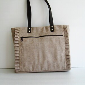 K&ouml;nnte beinhalten: Beige Tragetasche mit dunkelbraunen Ledergriffen und einer Rei&szlig;verschlusstasche vorne. Die Tasche hat einen dekorativen R&uuml;schenrand an den Seiten. Geeignet f&uuml;r den t&auml;glichen Gebrauch. Ma&szlig;e in Zentimetern.