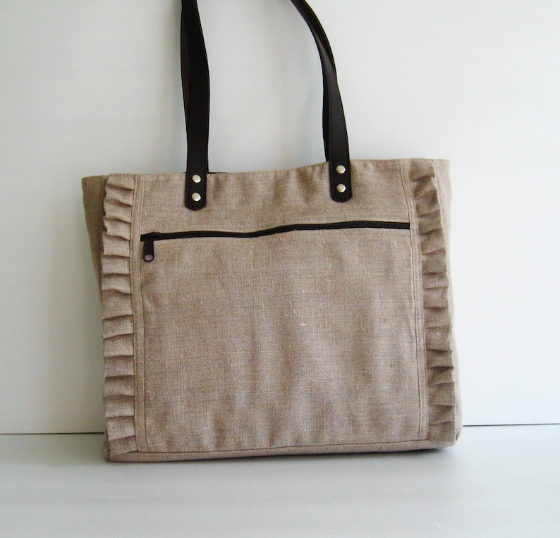 K&ouml;nnte beinhalten: Beige Tragetasche mit dunkelbraunen Ledergriffen und einer Rei&szlig;verschlusstasche vorne. Die Tasche hat einen dekorativen R&uuml;schenrand an den Seiten. Geeignet f&uuml;r den t&auml;glichen Gebrauch. Ma&szlig;e in Zentimetern.