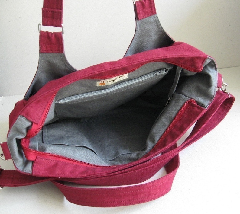 May include: A burgundy tote bag with gray lining and multiple pockets. The bag has a zippered main compartment and a long, adjustable shoulder strap. The interior features a small label with the text "Tippy Thai".