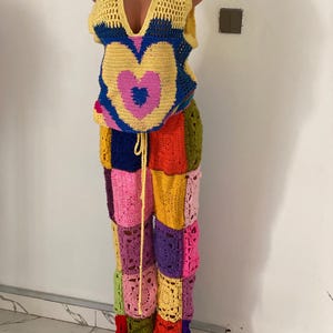 May include: A handmade crochet outfit. The top is yellow with a deep V-neck and a heart design in pink and blue. The patchwork trousers feature various colours. The outfit is displayed on a mannequin.