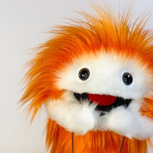 Puede incluir: Una marioneta caprichosa naranja y blanca con una textura peluda. La marioneta tiene grandes ojos negros, una boca roja y manos blancas. El pelaje naranja de la marioneta es puntiagudo, lo que le da una apariencia juguetona. La marioneta está diseñada para ser sostenida con dos varillas metálicas.