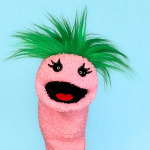 May include: A pink sock puppet with a smiling face, black eyes, and green hair. The puppet's mouth is open, revealing a red tongue. The background is a solid light blue.