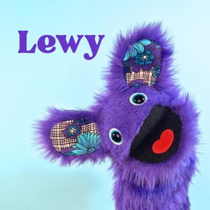 Puede incluir: Una criatura de peluche morada con el nombre "Lewy" en texto morado. La criatura tiene ojos grandes y redondos, una boca negra con un corazón rojo y tela estampada en las orejas. El fondo es azul claro.