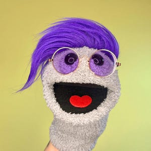 May include: A gray sock puppet with a purple wig and round, gold-rimmed glasses with purple lenses. The puppet has a black mouth with a red heart-shaped tongue. The background is a light green color.