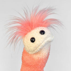 May include: A handmade sock puppet in pink and white. It features fluffy pink 'hair', two black button eyes, and a body that fades from pink to white. A whimsical, handcrafted item.