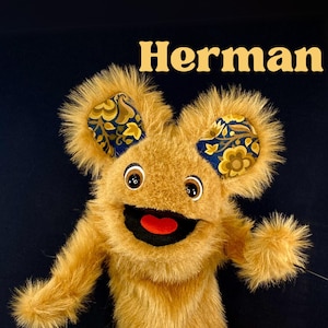 May include: A plush puppet with a tan fur body, large eyes, and a red mouth. The puppet's ears are adorned with patterned fabric in shades of blue, gold, and brown. The name "Herman" is written in gold letters above the puppet.