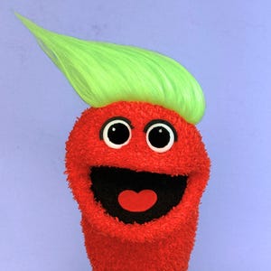 May include: A red fuzzy puppet with a bright green synthetic hair tuft, large white eyes with black pupils, and a wide open mouth featuring a red tongue. The puppet has a textured, plush surface and is set against a solid light blue background.