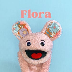 Könnte beinhalten: Eine rosa Plüsch-Handpuppe namens Flora mit einem strukturierten, lockigen Stoffkörper. Sie hat große Ohren mit einem bunten Blumenmuster in Orange, Blau und Weiß. Die Puppe hat schwarze Augen mit weißen Highlights und einen breiten Mund mit einer roten Zunge.