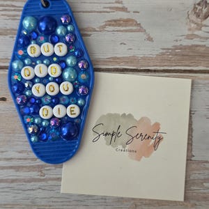 May include: A blue motel-style keychain decorated with various shades of blue beads, rhinestones, and white letter beads spelling out the phrase BUT DID YOU DIE. The keychain includes a silver metal ring and is displayed next to a square card.