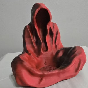以下が含まれることがあります： 座った姿勢のフード付きの赤い彫刻。滑らかで流れるようなローブと深いフードがあり、顔の特徴は見えません。彫刻は白い表面にあります。