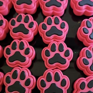 Pode incluir: Um close-up de vários objetos rosa em forma de pata, cada um com uma pegada preta. Os objetos estão dispostos em uma grade, com as pegadas claramente definidas. O material parece ser plástico ou uma substância semelhante.