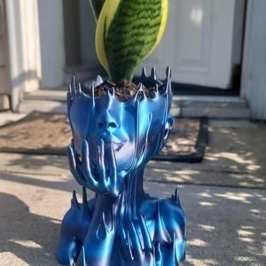 May include: A blue decorative planter in the shape of a human head, with hands supporting the face. The planter holds a snake plant with green and yellow leaves. The planter is made of a shiny, metallic-looking material.