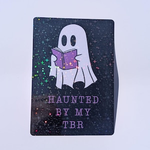 May include: A rectangular sticker with a black holographic background featuring a white ghost reading a purple book. The text HAUNTED BY MY TBR is printed in purple below the ghost. The background has a shimmering star pattern throughout the design.