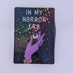 May include: A rectangular sticker with a black background featuring a holographic star pattern. The text IN MY HORROR ERA is printed in white. A purple hand with long nails holds a white eyeball, and a gold-colored bracelet is on the wrist.