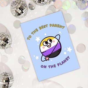 May include: A light blue greeting card with the text "TO THE BEST PARENT ON THE PLANET". The card features a cartoon planet with the non-binary pride flag colors. Silver disco balls and confetti are in the background.