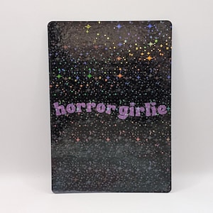 Peut inclure: Un autocollant rectangulaire avec un fond holographique noir présentant un motif dense de petites étoiles et d'étincelles multicolores. Le texte "horror girlie" est imprimé au centre dans une police violette claire, arrondie et bulleuse.
