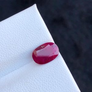 Puede incluir: Una gema de rubí facetada, de forma ovalada y color rojo intenso. La gema está colocada sobre una superficie blanca, con un fondo oscuro. Los rubíes son piedras preciosas, a menudo utilizadas en joyería. Mide aprox. 1 cm.