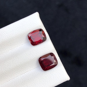 Könnte beinhalten: Zwei kissenförmige, tiefrote Edelsteine werden auf einer weißen Oberfläche vor einem schwarzen Hintergrund präsentiert. Die Edelsteine haben eine satte, gesättigte Farbe und eine polierte, reflektierende Oberfläche, die ihre Klarheit und Brillanz zeigt. Die Edelsteine sind wahrscheinlich für Schmuck.