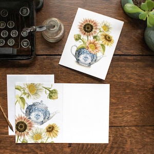 May include: Watercolor art print of sunflowers in a blue and white teapot. The image is on a white card, with a matching card and envelope. The scene is set on a dark wood surface with a vintage typewriter and plants.