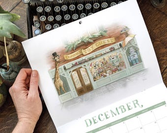 Puede incluir: Una página de calendario de pared para diciembre con una ilustración en acuarela de una tienda de juguetes con el letrero Duncan's Toy Chest. La tienda tiene una fachada verde claro, una jirafa con sombrero de copa y un escaparate con juguetes.