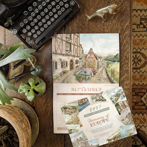 Puede incluir: Un calendario de pared de 2027 titulado Dreaming of Europe con ilustraciones en acuarela de pueblos europeos. El calendario muestra el mes de septiembre con una escena de casas con entramado de madera y un camino de piedra, junto a una máquina de escribir.