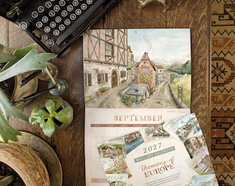 Op de afbeelding: Een wandkalender voor 2027 getiteld Dreaming of Europe met aquarelillustraties van Europese dorpen. De kalender toont de maand september met een tafereel van vakwerkhuizen en een stenen pad, naast een vintage typemachine.