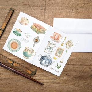 May include: Watercolor illustrations of teacups, teapots, and tea accessories on a white paper, with the text "Always & Infinity". A white envelope, a wooden ruler, and a pen are also present on a wooden surface.