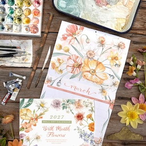Puede incluir: Un calendario de pared de 2027 titulado Birth Month Flowers con ilustraciones en acuarela de diversas flores. El calendario se muestra junto a una paleta de pintura, tubos de pintura, pinceles y flores frescas sobre una superficie de madera.