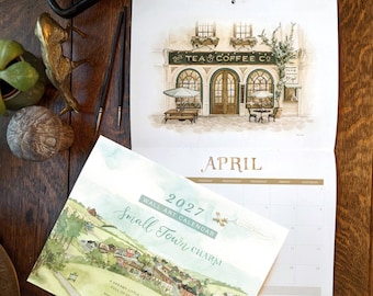 Puede incluir: Un calendario de pared de 2027 titulado Small Town Charm con ilustraciones en acuarela de un paisaje urbano y la fachada de una tienda de té y café. El calendario muestra el mes de abril con un diseño de cuadrícula y dibujos arquitectónicos.