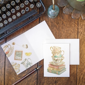 Könnte beinhalten: Ein Vintage-Themenbild mit Schreibwaren und einer Schreibmaschine. Zwei Grußkarten zeigen Teetassen, Teekannen und Bücher. Ein Stift und ein Tintenfass sind ebenfalls vorhanden. Die Szene ist auf einer Holzoberfläche angeordnet, was Nostalgie auslöst.
