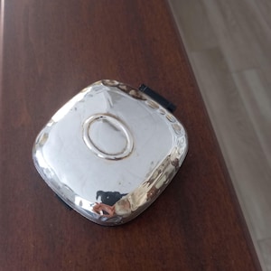 Może przedstawiać: Srebrna, kwadratowa szkatułka na drobiazgi z lekko wypukłym, owalnym wzorem w środku. Szkatułka ma klapkę na zawiasach i mały, czarny zatrzask.