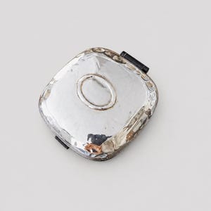May include: A square silver compact mirror with a hammered, textured surface and rounded corners. The metallic case features a central oval indentation and a black hinge mechanism visible on the side, reflecting light across its polished finish.