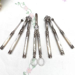 May include: A set of antique silver-toned serving utensils, including nutcrackers, picks, and scissors. The handles are inlaid with mother-of-pearl, and the set is arranged on a white surface with floral embroidery. The utensils have ornate detailing.