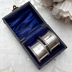 May include: A pair of polished silver napkin rings with engraved monogram details, presented inside a rectangular black hinged box. The interior of the box is lined with deep blue satin and velvet fabric, featuring a gold-toned metal latch closure.