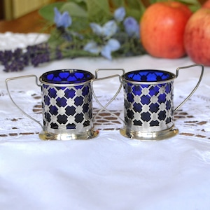 May include: Two silver-plated cups with blue glass inserts and a lattice design. The cups have handles and are sitting on a white lace tablecloth.
