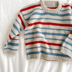 Pode incluir: Um su&eacute;ter de malha com listras horizontais vermelhas, azul claro e bege. O su&eacute;ter tem mangas compridas e gola redonda. O tecido parece ser uma malha grossa, e a pe&ccedil;a de roupa est&aacute; sobre uma superf&iacute;cie branca.