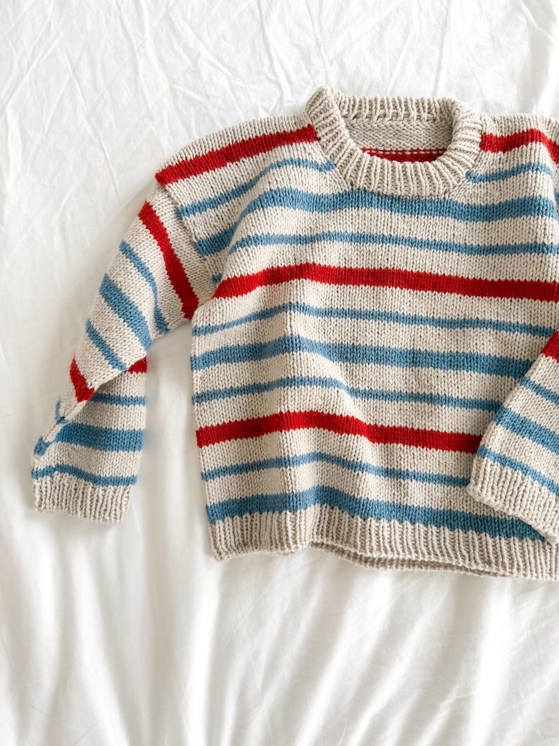 Pode incluir: Um su&eacute;ter de malha com listras horizontais vermelhas, azul claro e bege. O su&eacute;ter tem mangas compridas e gola redonda. O tecido parece ser uma malha grossa, e a pe&ccedil;a de roupa est&aacute; sobre uma superf&iacute;cie branca.