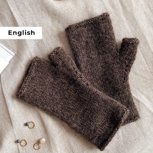 Op de afbeelding: Een paar bruine vingerloze handschoenen, gebreid met een getextureerde steek. De handschoenen zijn gemaakt van een donkerbruin garen en worden gepresenteerd op een neutraal gekleurd linnen oppervlak. De afbeelding bevat ook breinaalden en kleine gouden oorbellen.