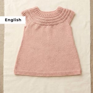 Peut inclure: Robe tricotée rose clair avec un col texturé et des manches courtes. La robe est faite d'un fil doux et texturé et est présentée sur une surface blanche. Le design est simple et classique.