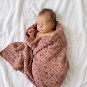 May include: A dusty rose knitted baby blanket featuring a delicate eyelet lace pattern and a garter stitch border. The blanket is wrapped around a sleeping infant, showcasing the soft texture and intricate openwork design of the fabric.