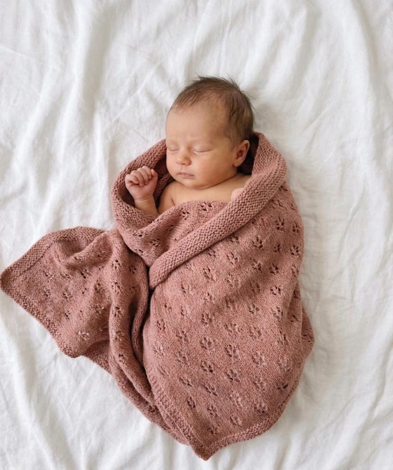 May include: A dusty rose knitted baby blanket featuring a delicate eyelet lace pattern and a garter stitch border. The blanket is wrapped around a sleeping infant, showcasing the soft texture and intricate openwork design of the fabric.