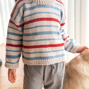 Pode incluir: Um su&eacute;ter de malha com listras horizontais vermelhas, azul claro e bege. O su&eacute;ter tem gola redonda e mangas compridas. As mangas e a bainha inferior t&ecirc;m um design canelado. O su&eacute;ter &eacute; usado com cal&ccedil;as cinzentas.