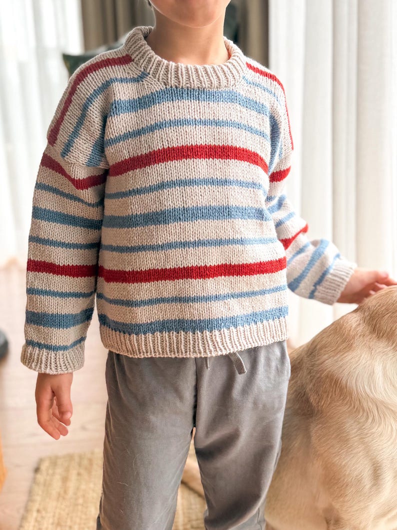 Pode incluir: Um su&eacute;ter de malha com listras horizontais vermelhas, azul claro e bege. O su&eacute;ter tem gola redonda e mangas compridas. As mangas e a bainha inferior t&ecirc;m um design canelado. O su&eacute;ter &eacute; usado com cal&ccedil;as cinzentas.