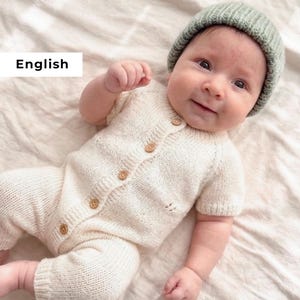 Puede incluir: Un bebé con un pelele de punto color crema con botones de madera y un gorro a juego verde salvia. El pelele tiene mangas cortas y pantalón. El bebé está sobre una sábana crema.